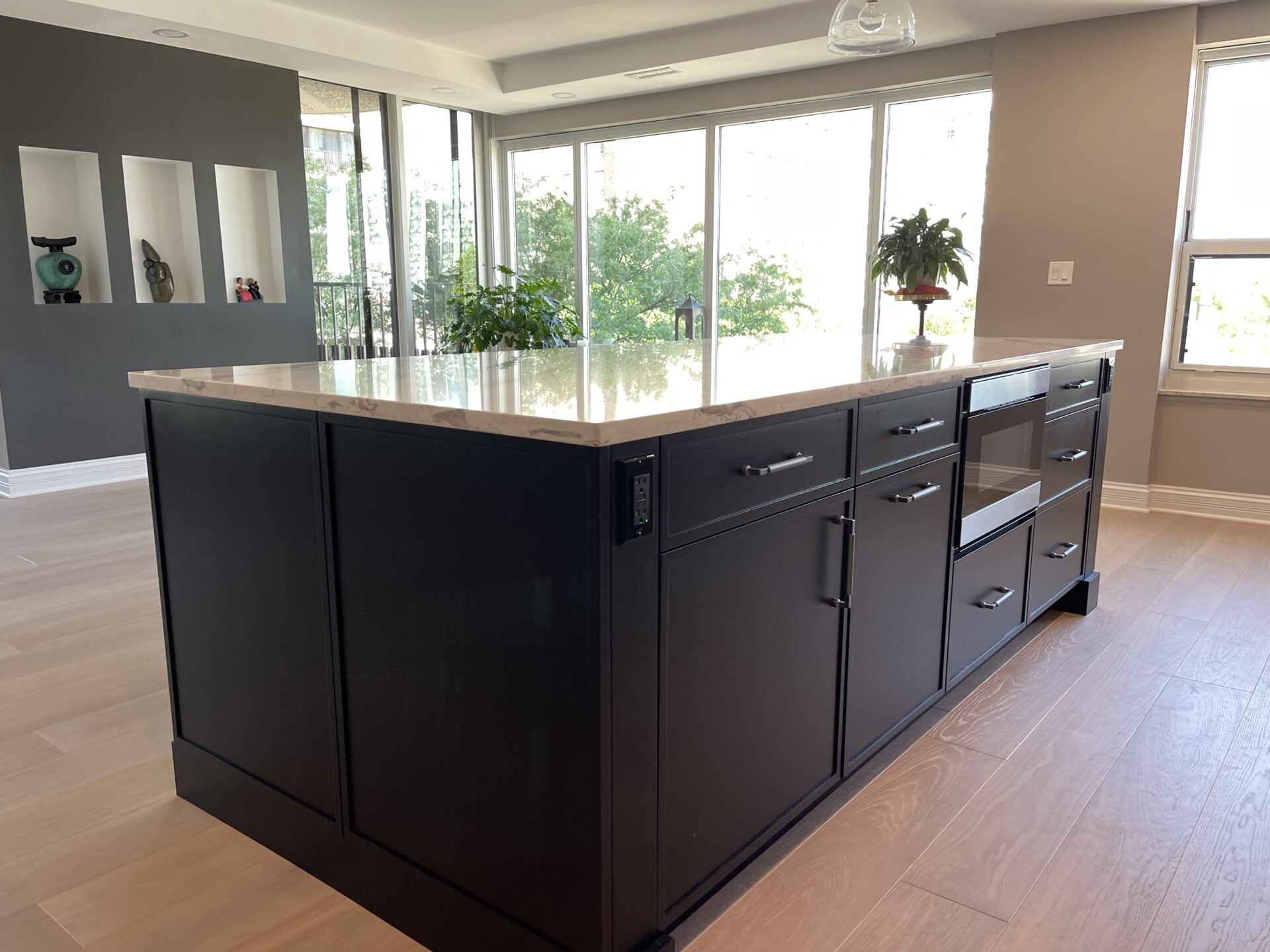 Large black kitchen island with white countertop in foreground, windows showing greenery in the background
