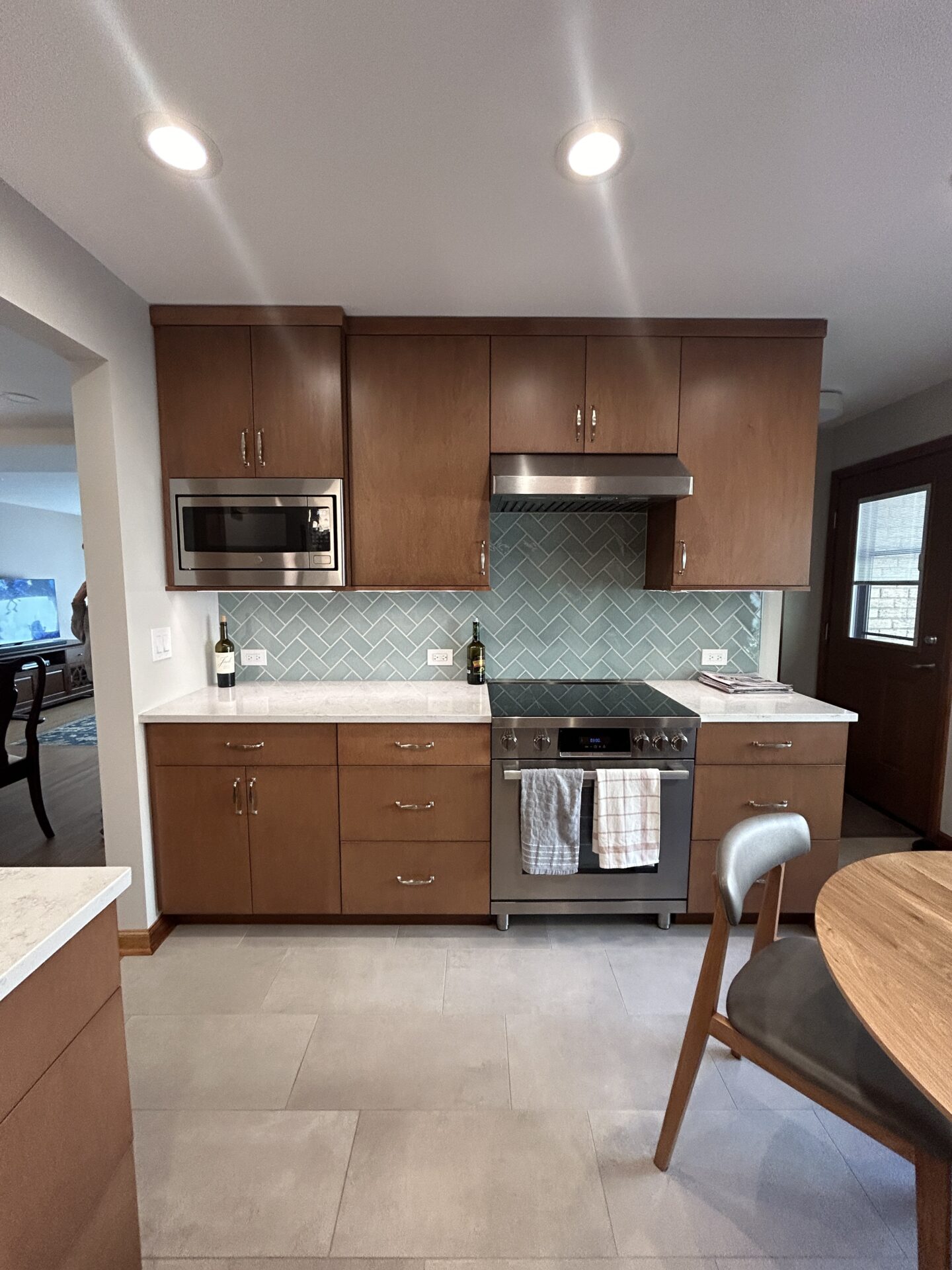Maple cabinets in kitchen with herringbone blue green backsplash, white countertops, peninsula to left, table to right, stainless steel range, hood and above counter microwave along cabinetry wall